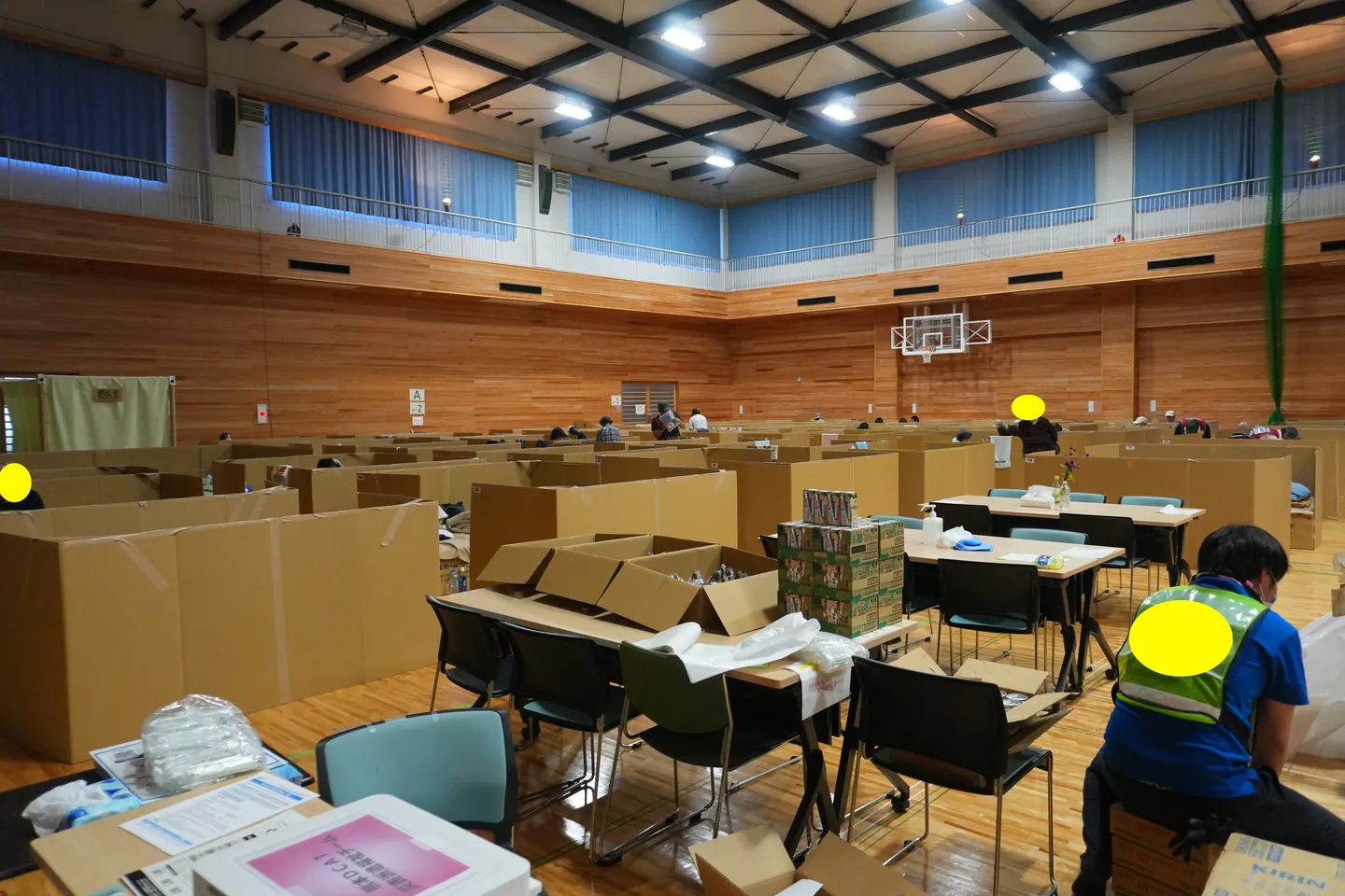 体育館の床に間仕切りや生活用品が並び、支援スタッフが活動する避難所の様子